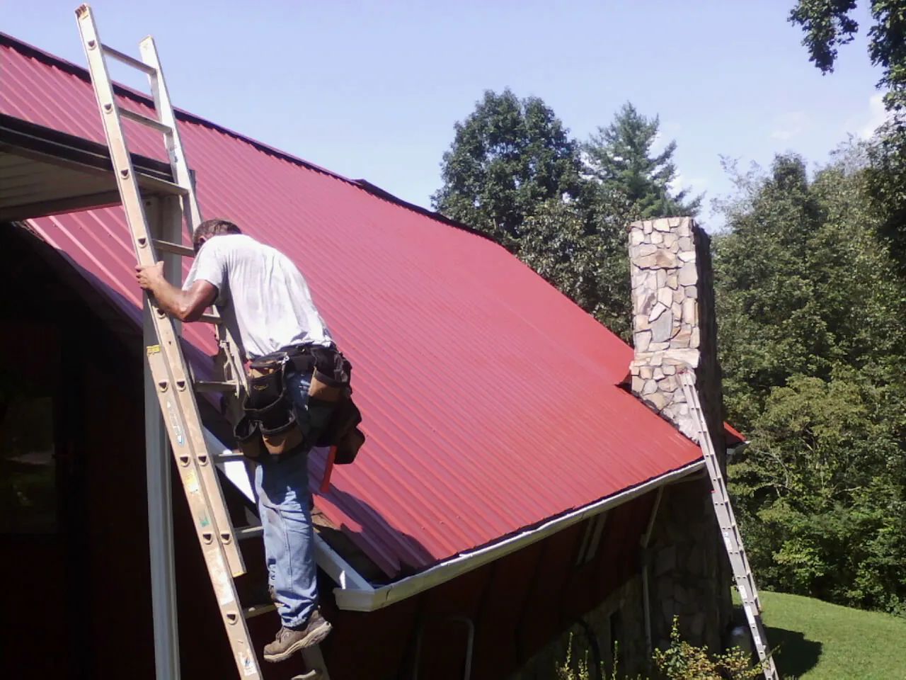 Roofer on a ladder beside a red metal roof and stone chimney — Soffit & Fascia Repair by Roofing Brothers in Lincoln
